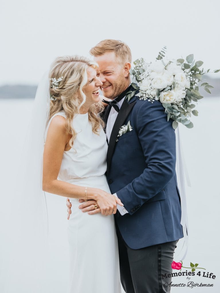 Brud och brudgummi med blomsterbukett vid vattnet.